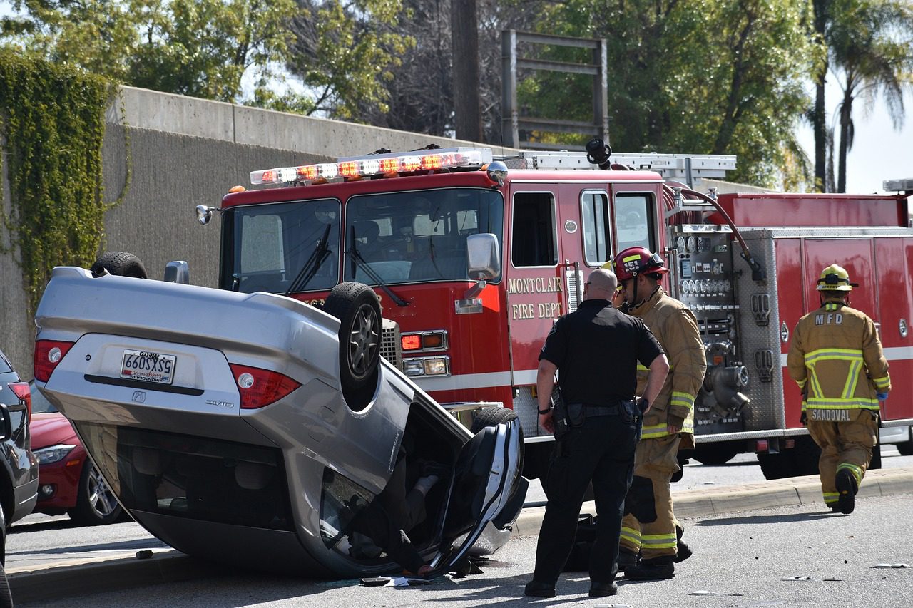 A policeman wearing a black uniform stands beside a firefighter looking at the silver car near the red fire truck