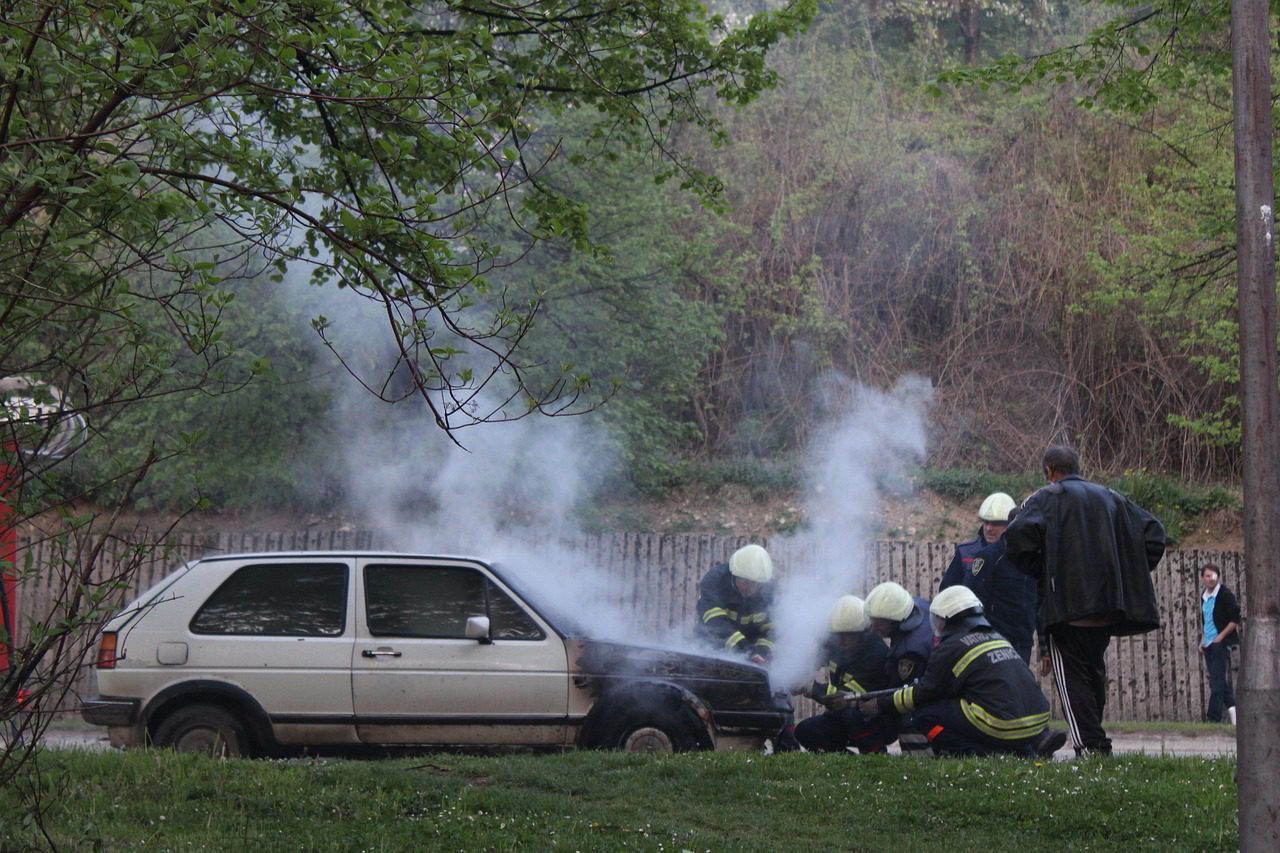 Five firefighters are extinguishing the fire on a white car near a brown wooden fence on an empty street