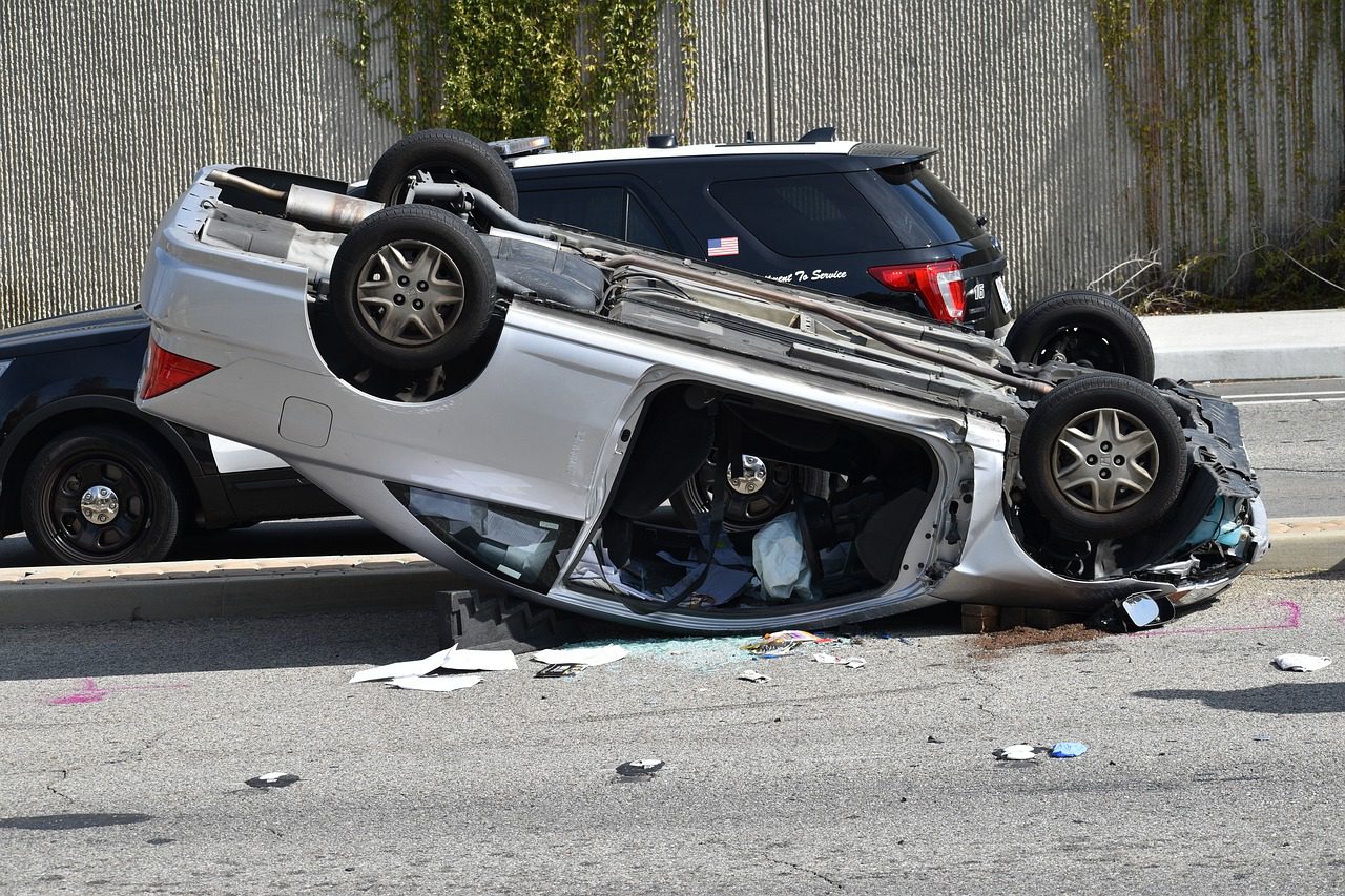 A white car is flipped upside down beside a black SUV near a gray wall