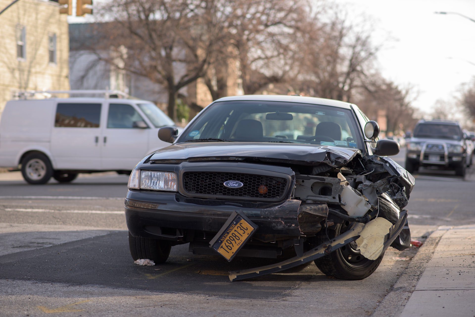 A wrecked black Ford car has a broken bumper and an almost detached yellow plate number near a sidewalk