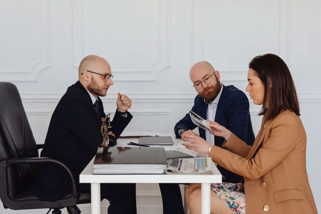 Woman in beige blazer holding photos while two of her lawyers are listening to her