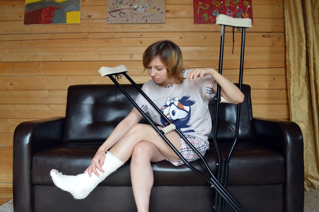 Woman sitting down on a black leather couch while holding on to her crutches and leg that had a cast