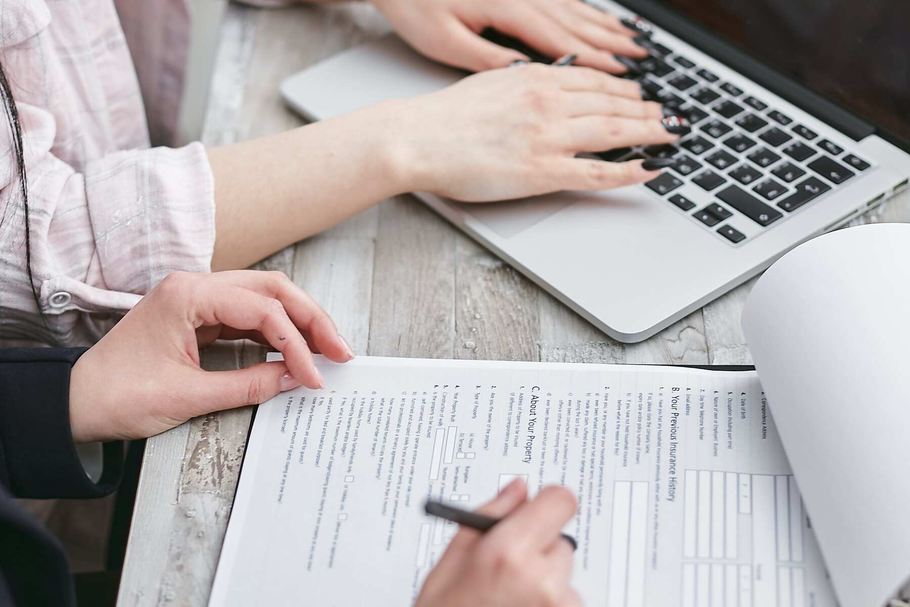 A woman typing on the laptop while another woman holds a pen and a form